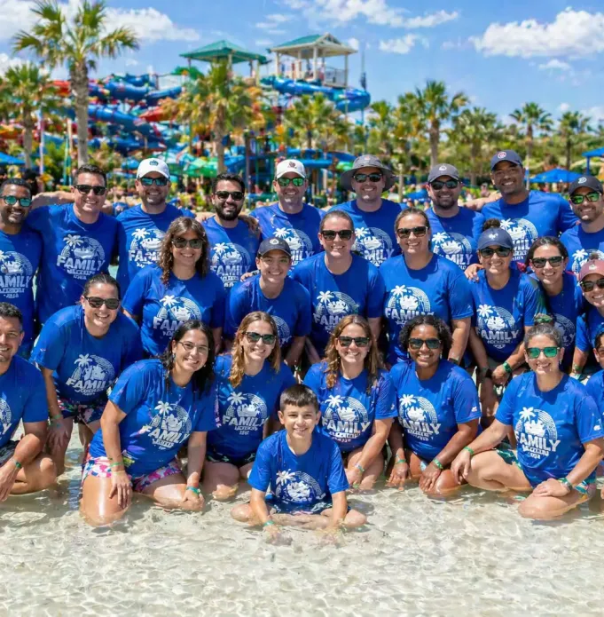 Group of about 25 people wearing matching blue shirts posing together in a shallow waterpark pool.