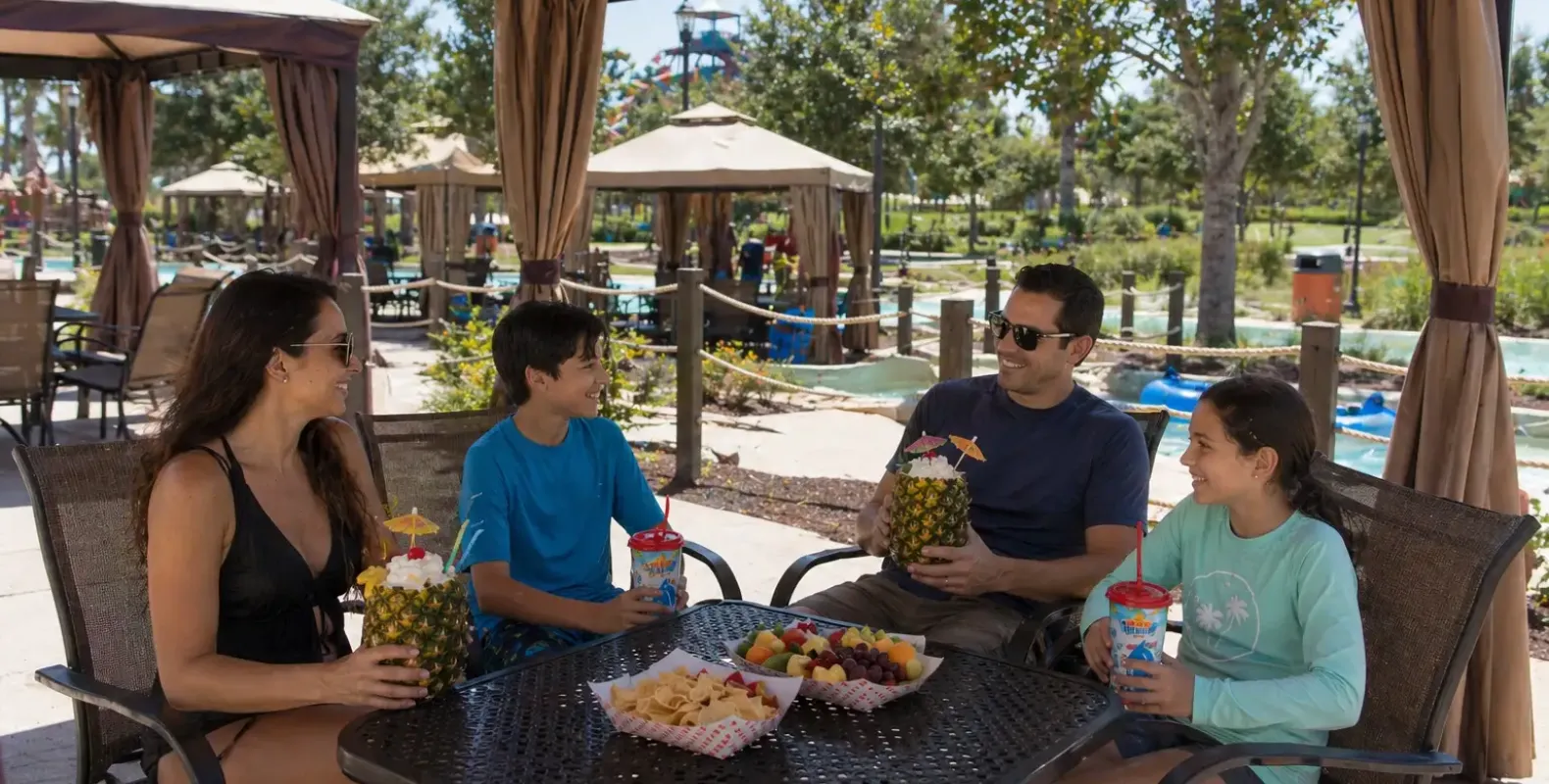Family of four relaxing under a shaded waterpark cabana with snacks and tropical drinks beside a lazy river.