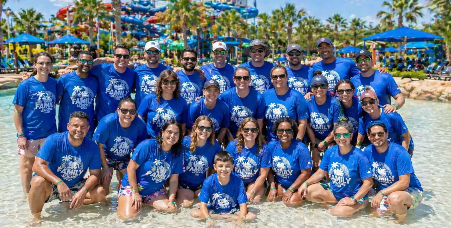 Group of about 25 people wearing matching blue shirts posing together in a shallow waterpark pool.