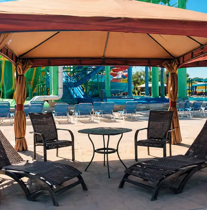 Shaded waterpark cabana with two lounge chairs, two upright chairs, and a small table near colorful slides and poolside seating.