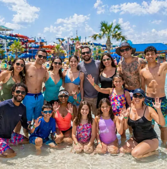 Small group of adults and children smiling together in a shallow waterpark pool on a sunny day.